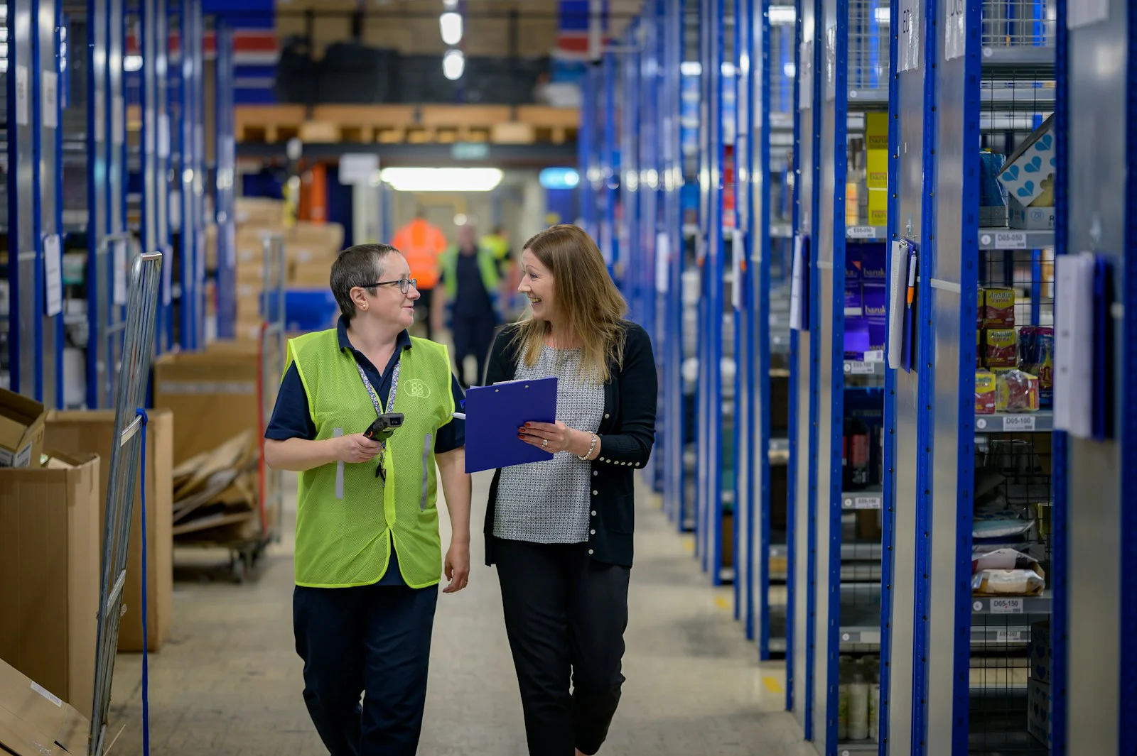 Two colleagues talking in a warehouse aisle, one in a hi-vis vest